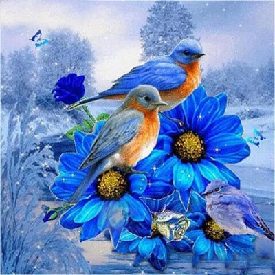 A decorative floral scene featuring two bluebirds perched on large bright blue flowers with yellow centers, set against a snowy blue landscape with trees and small butterflies. Birds in Blue - Diamond Painting