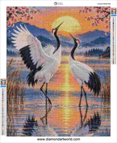 A diamond painting artwork featuring two white cranes with black wings standing in shallow water at sunset, with pink blossoms, mountains, and a glowing sun in the background. The image includes a brand header at the top and color swatch bars on the sides. Cranes - Diamond Painting